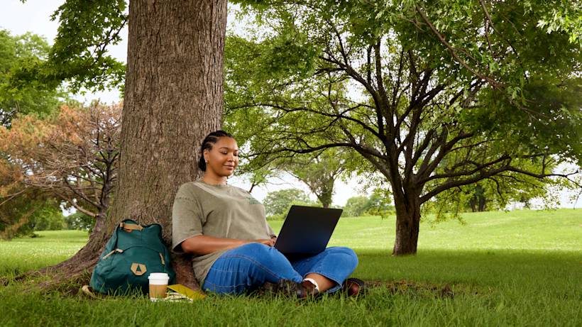 Person relaxing under a large tree in a park, using laptop while leaning against trunk with backpack and coffee nearby