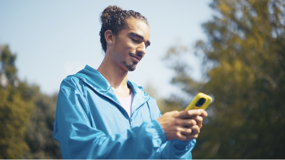 Person in bright blue jacket looking down at yellow smartphone outdoors on sunny day with trees in background