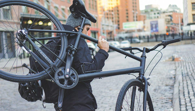 A man in a black jacket and gray beanie holds a black bike over his shoulder as he looks down a cobblestone street lined with shops.